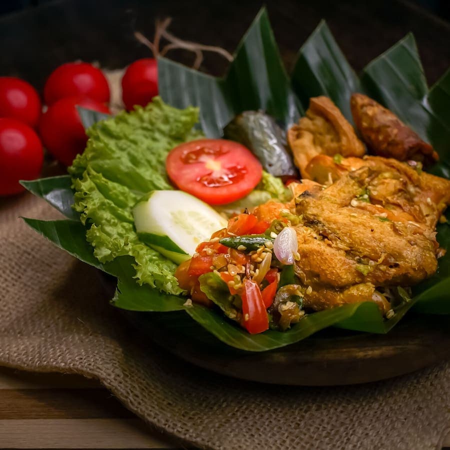 Ayam penyet dengan sambal pecak, lalapan, dan irisan tomat di atas daun pisang